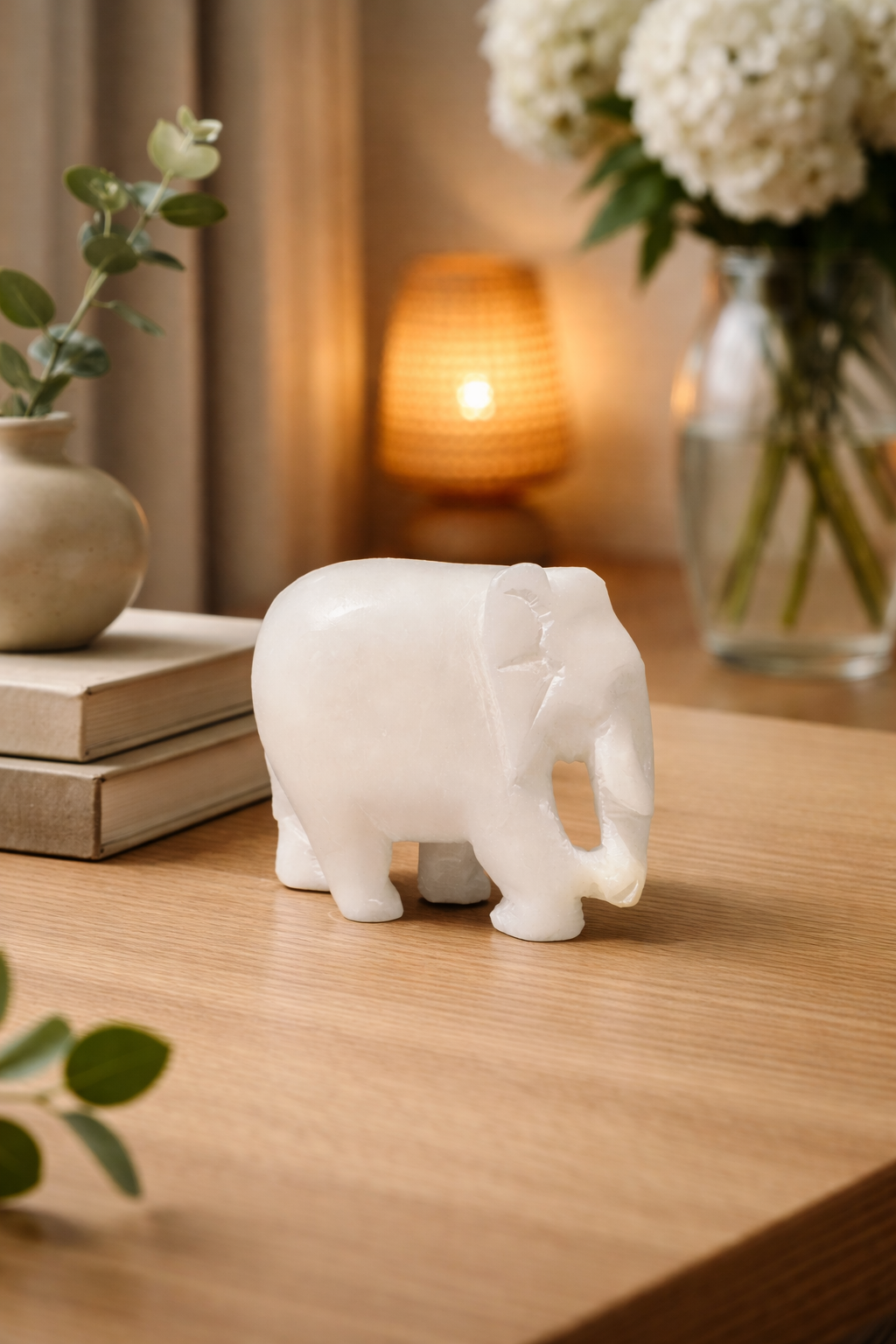 Polished white marble elephant sculpture on wooden table with warm lamp and flowers in background