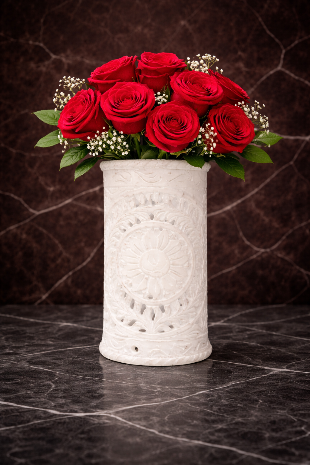 White marble carved vase with intricate mandala detailing holding a bouquet of red roses and baby’s breath on a dark marble surface.