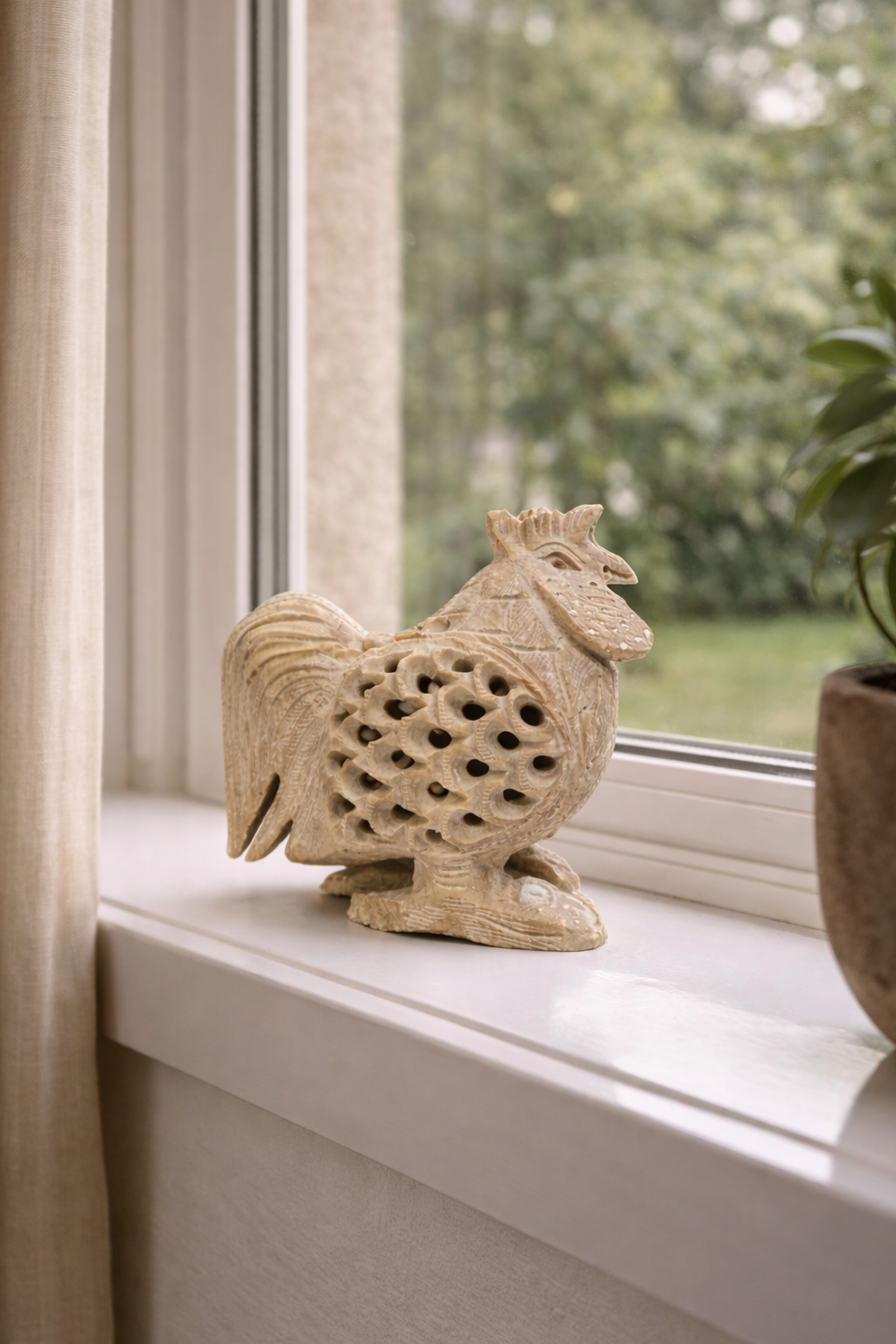 Hand-carved soapstone rooster sculpture displayed on windowsill with greenery outside