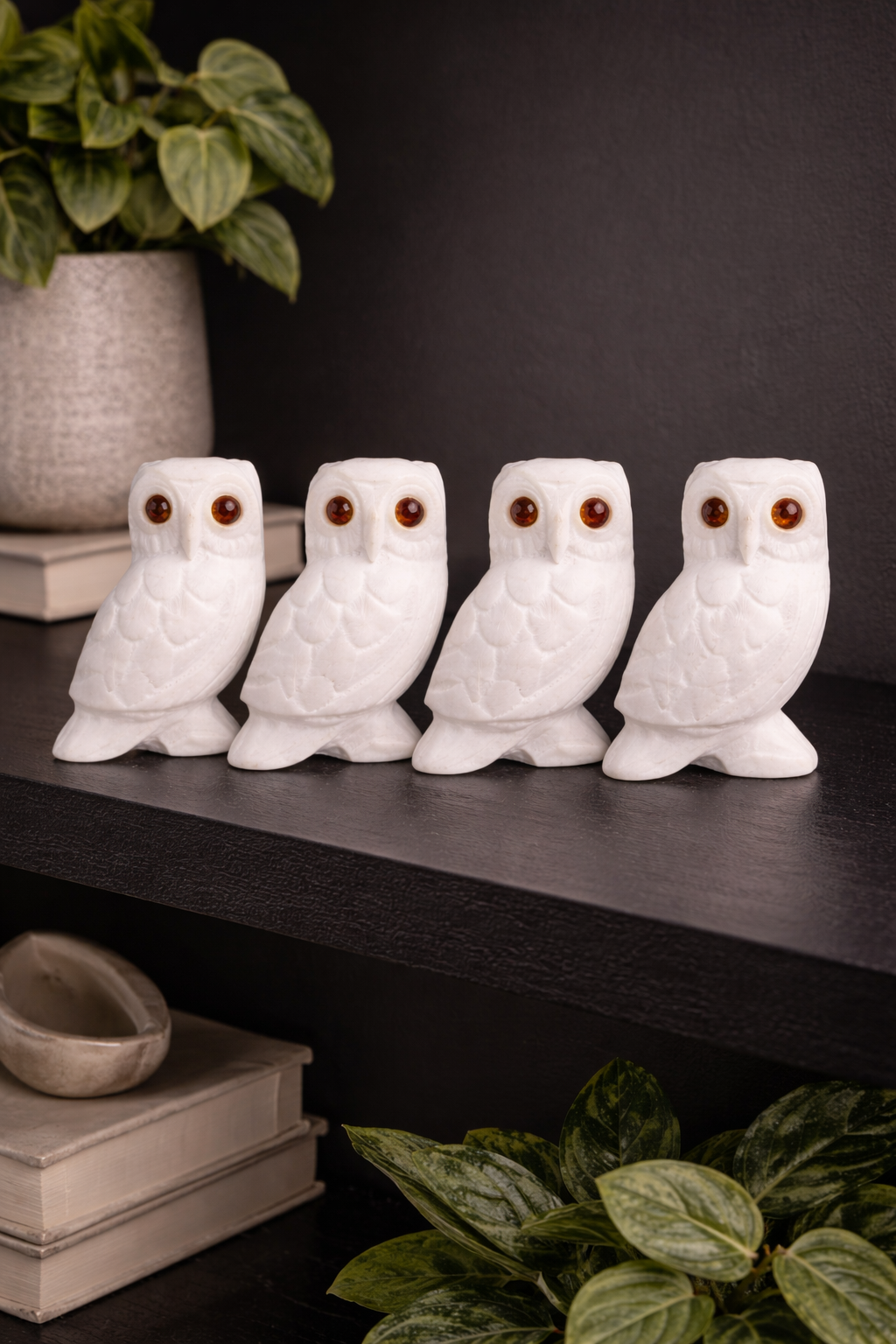 Four white marble owl figurines with red stone eyes displayed on dark shelf with plants in modern home