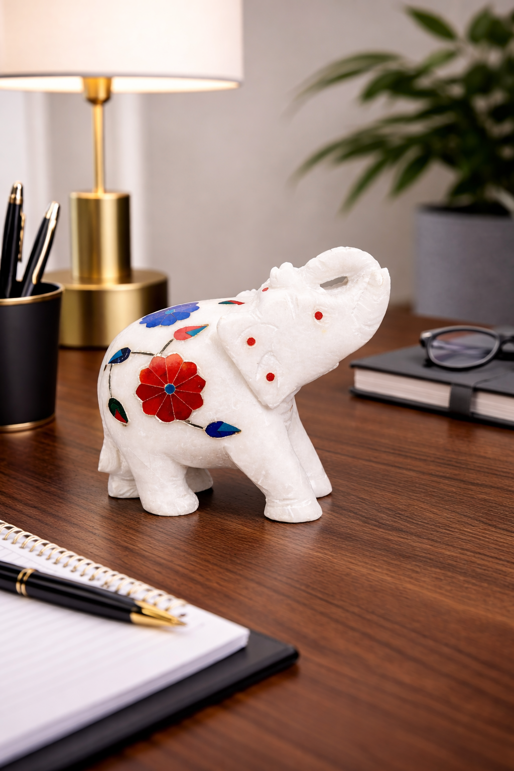 Hand-carved white marble elephant figurine with colourful floral inlay placed on an office desk with lamp and notebook – luxury decorative sculpture.